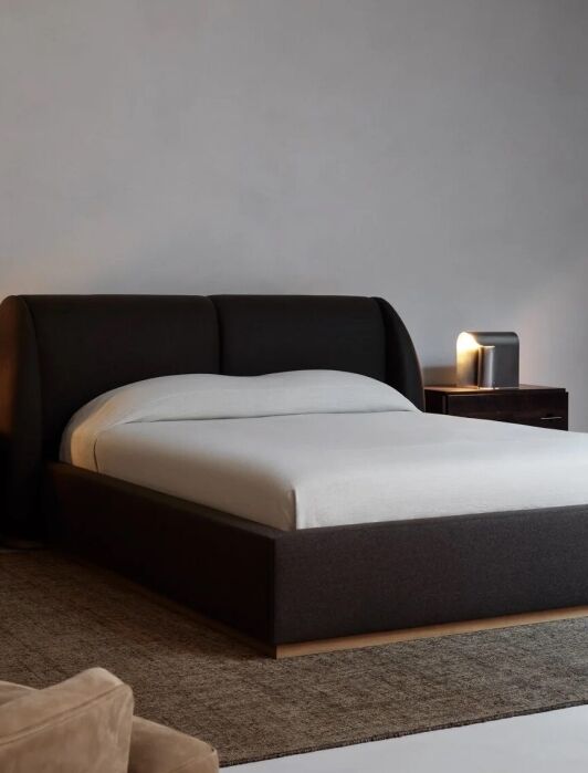 Minimalist bedroom with a dark upholstered bed, two wooden nightstands with lamps, a wooden bench, and a large neutral rug on a concrete floor. Walls are plain and light gray.