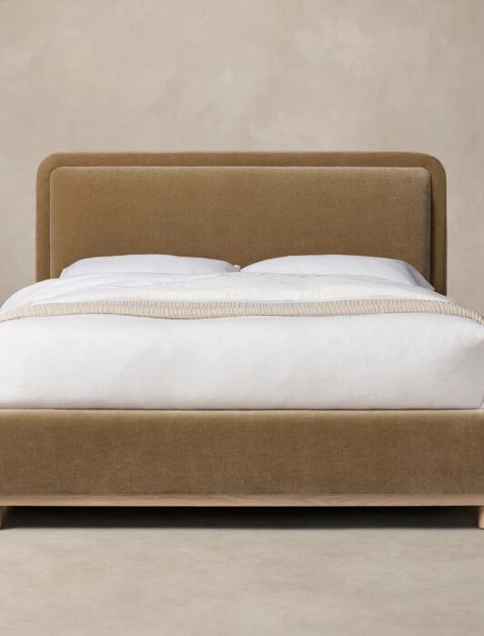 Bed with a tan upholstered frame, white bedding, and light wooden legs, positioned against a beige background.