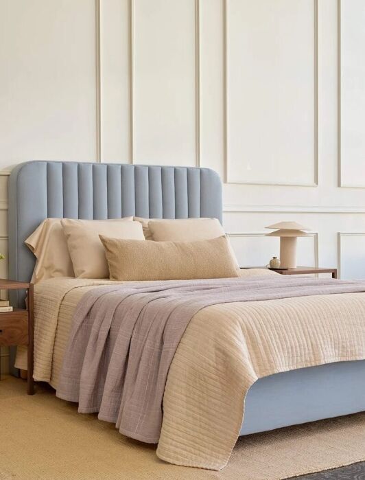 A neatly made bed with a light blue upholstered headboard, flanked by two wooden nightstands, sits in a bright, modern bedroom with large windows and paneled walls.