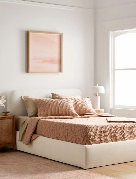 A neatly made bed with beige bedding and pillows, a wooden nightstand with flowers, a table lamp, a window, and a framed abstract art piece above the bed.