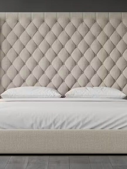 A beige upholstered bed with a tall, diamond-tufted headboard, white pillows, and a white mattress, set against a dark gray wall and wooden floor.