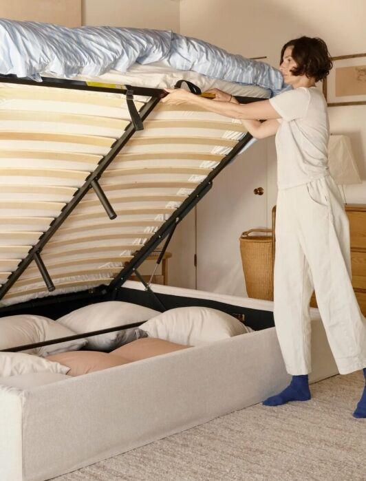 A person in white clothes lifts the storage compartment of a beige bed frame, revealing pillows inside. The room has a wooden dresser, artwork, and neutral decor.