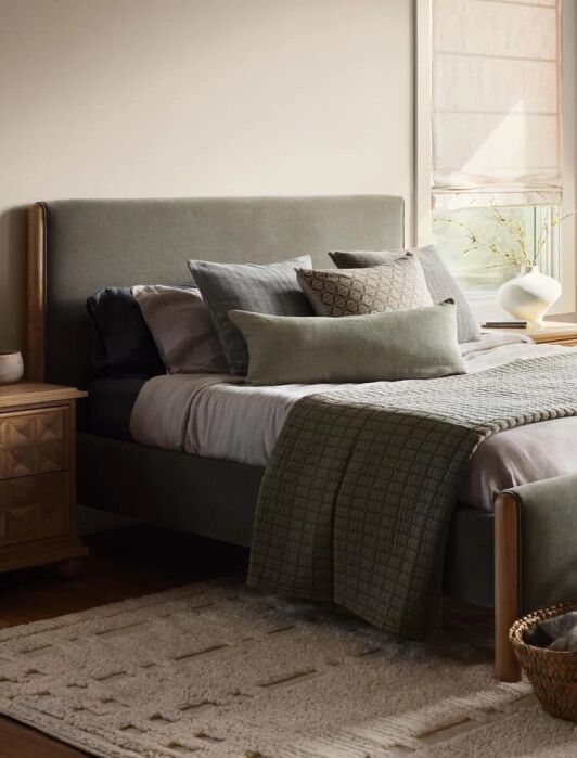 A neatly made bed with gray-green bedding, decorative pillows, a nightstand with a vase, woven baskets, and a window letting in natural light.