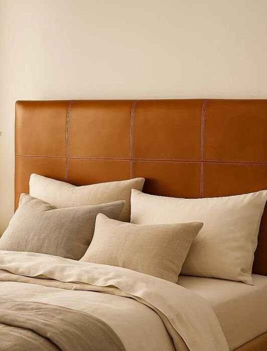 Bed with tan leather headboard, beige and cream bedding, and several pillows. A vase with dried grasses sits on a wooden nightstand against a neutral wall.