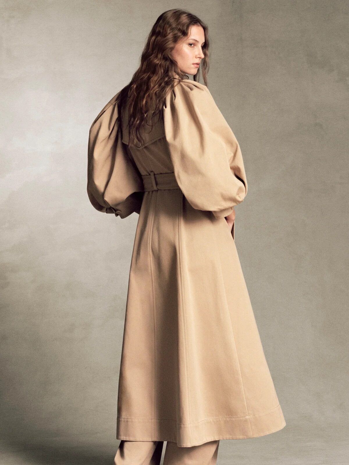 Person wearing a long, beige trench coat with voluminous sleeves and matching wide-leg pants, standing on a neutral background.