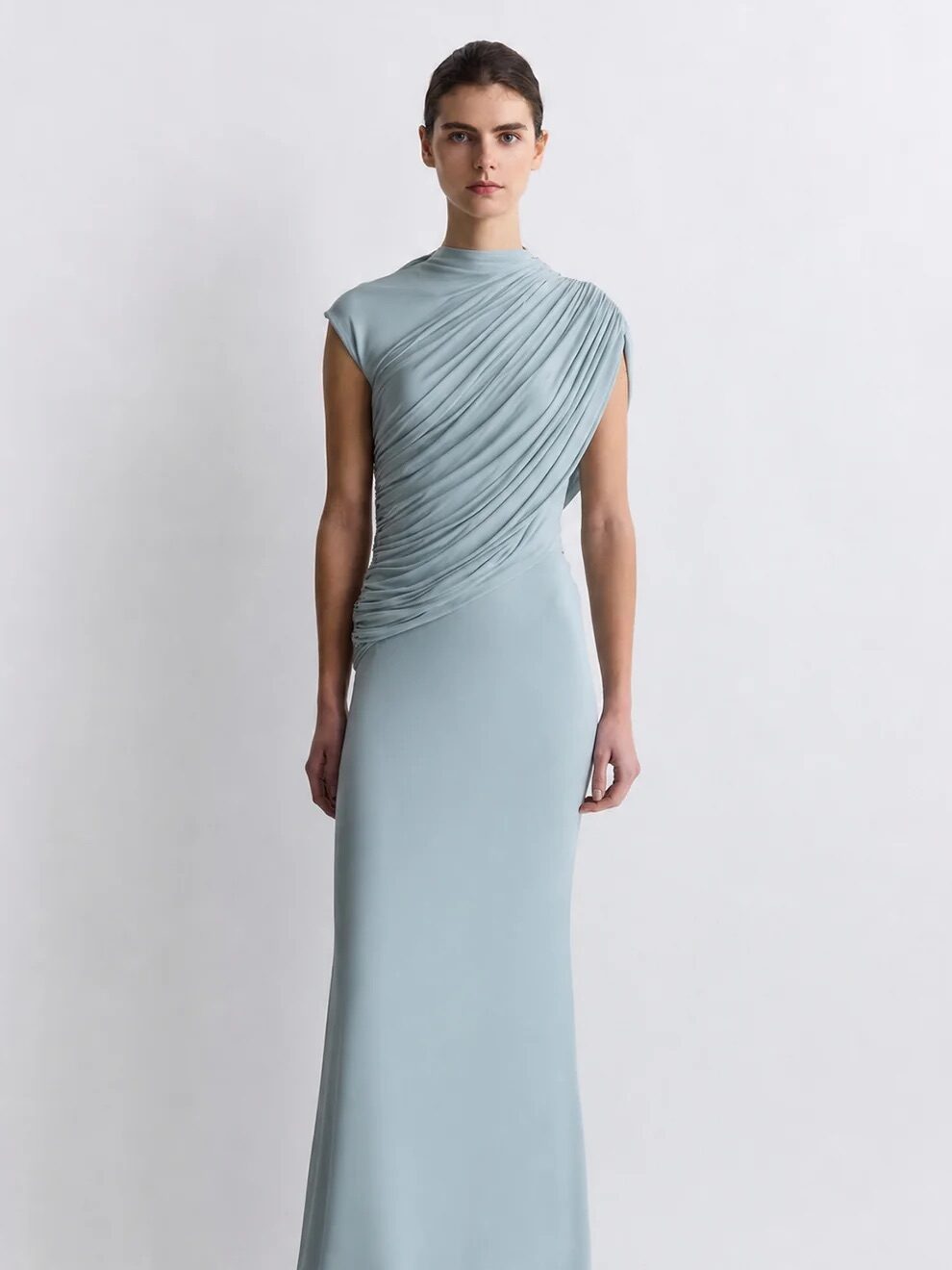 A person stands wearing a light blue, floor-length gown with draped fabric detail across the bodice against a plain light background.