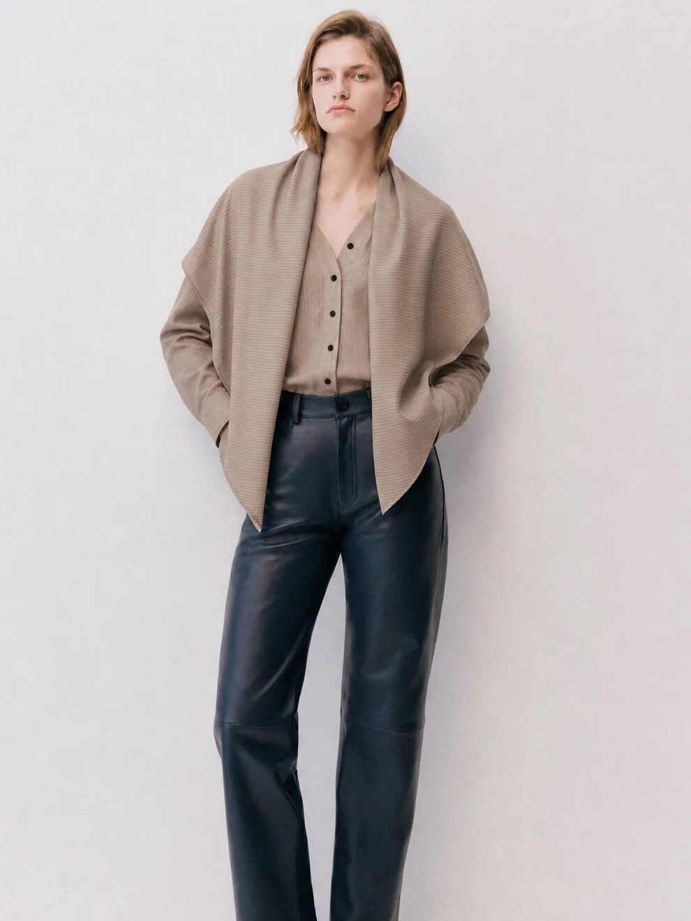 Person standing against a plain background, wearing a taupe cardigan over a beige buttoned shirt, black leather pants, and black sandals, with hands in pockets.