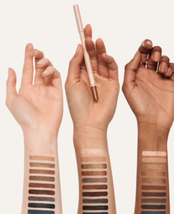Three arms of different skin tones display swatches of various makeup shades from light to dark, with the middle hand holding a dual-ended makeup pencil.