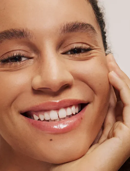 Close-up of a person smiling with clear skin, glossy lips, and a hand resting on their face against a neutral background.