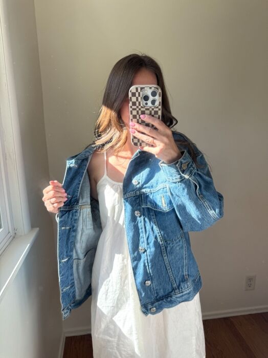 Person wearing a white dress and blue denim jacket stands indoors, taking a mirror selfie with a checkered phone case. Sunlight enters through a nearby window.