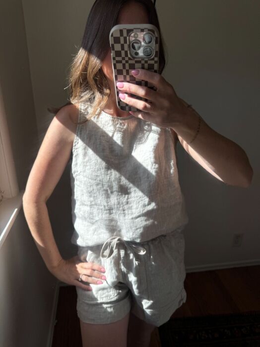 Person wearing light-colored sleeveless linen top and shorts stands indoors, holding a phone with a checkered case for a mirror selfie. Sunlight streams in from the left.