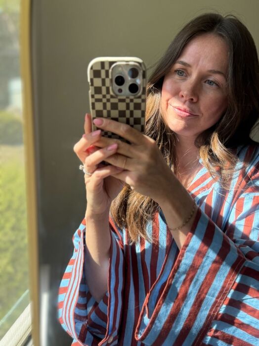 A woman in a blue and brown striped robe takes a selfie with a checker-patterned phone by a sunlit window.