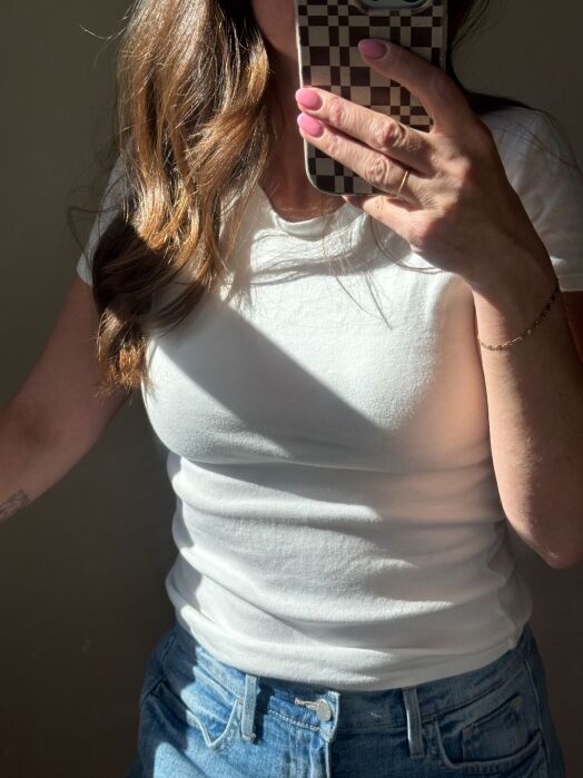 A person in a white t-shirt and blue jeans takes a mirror selfie using a phone with a checkered case. Sunlight highlights part of their torso and hair.