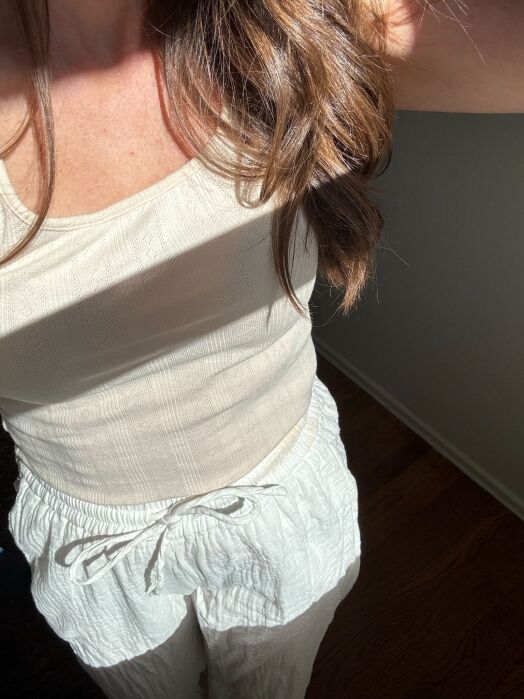 Woman wearing a cream-colored tank top and drawstring pants stands in sunlight, with her face out of frame and brown hair cascading over her shoulder.