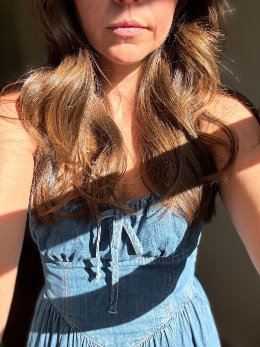 A woman wearing a blue denim dress is standing in sunlight, with her face partially visible and long wavy brown hair over her shoulders.