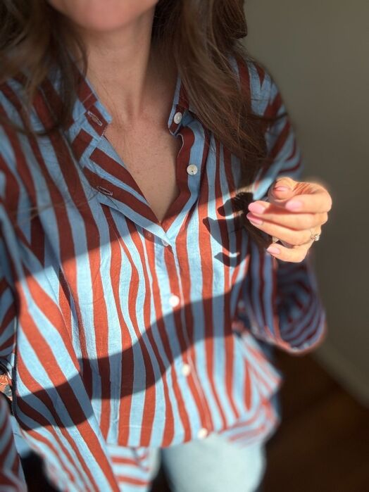A woman wearing a blue and brown striped button-up shirt stands in sunlight, holding something small in her hand. Her face is partially out of frame.