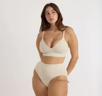 A woman stands against a plain background, wearing a matching light beige bralette and high-waisted underwear.