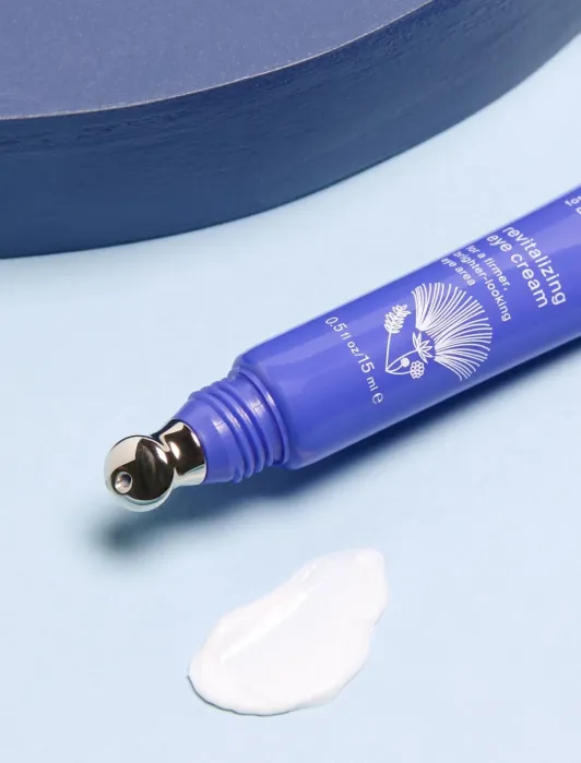 A blue tube of revitalizing eye cream with a metal applicator tip lies next to a dollop of cream on a light blue surface.