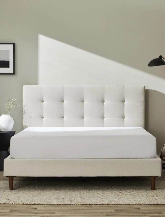 Minimalist bedroom with a beige upholstered bed, tufted headboard, side table with vase, modern lamp, chair, and framed wall art against a light gray wall.
