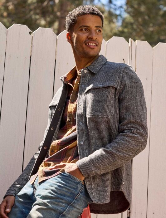 Man wearing a gray shirt jacket over a plaid shirt and jeans, sitting on a red stool in front of a white wooden fence outdoors.