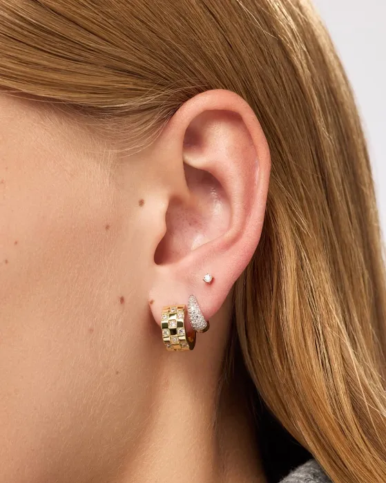 Close-up of a person’s ear with three earrings: a small stud in the upper lobe, a textured silver hoop, and a gold geometric hoop in the lower lobe. Light brown hair partially visible.