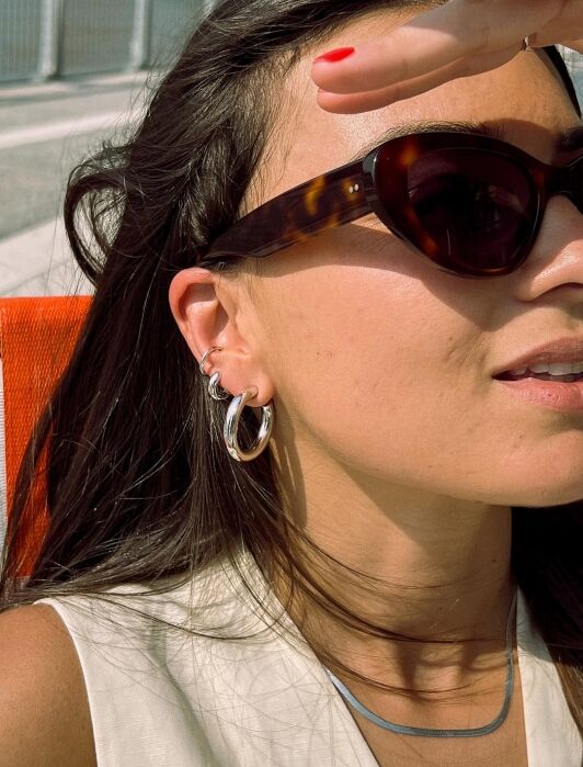 A person wearing dark sunglasses, silver hoop earrings, and a sleeveless top shields their eyes from the sun with their hand.