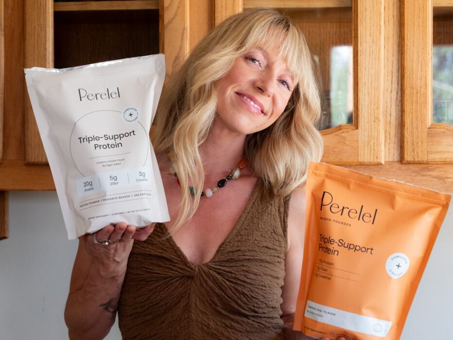 Smiling woman holding two bags of Perelel Triple-Support Protein powder, one white and one orange, standing in front of wooden cabinets.