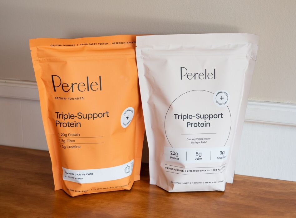 Two bags of Perelel Triple-Support Protein powder, one orange and one white, placed on a wooden surface against a white wall.