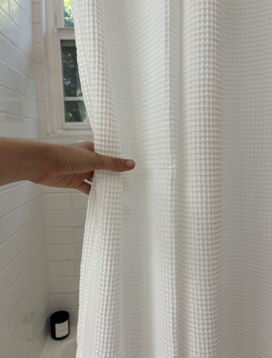 A hand pulls back a textured white shower curtain in a tiled bathroom with a small black container on the floor.
