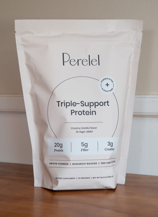A bag of Perelel Triple-Support Protein powder sits on a wooden surface. The front label highlights 20g protein, 5g fiber, 3g creatine, creamy vanilla flavor, and no sugar added.