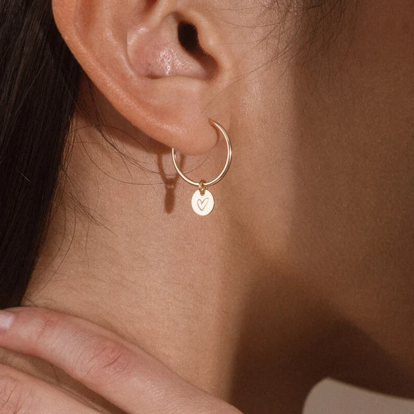 Close-up of a person’s ear wearing a gold hoop earring with a small round pendant engraved with a heart. A hand rests gently on the neck.