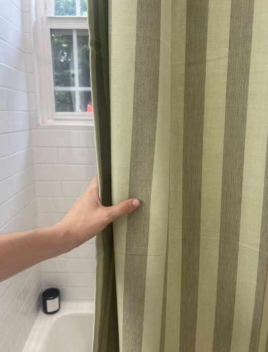 A hand holds an olive and beige striped shower curtain next to a white tiled wall, bathtub, and window.