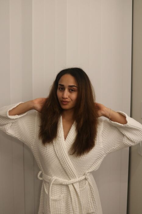 A person wearing a white bathrobe stands indoors, facing the camera with their hands raised to touch their hair, in front of vertical white blinds.