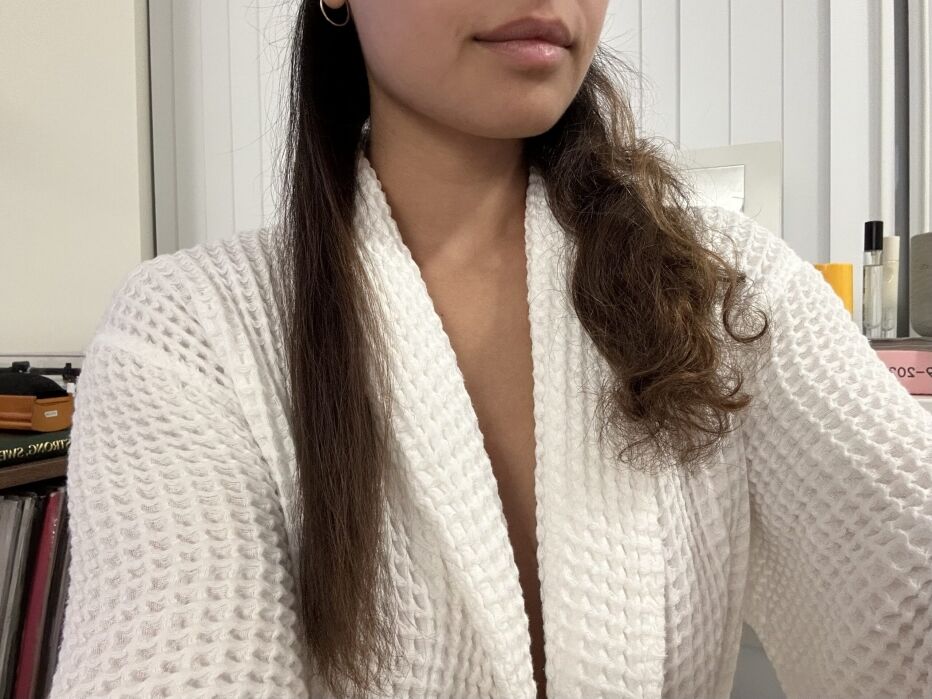 A person in a white robe shows their hair, with one half straight and the other half curly, against a background of shelves and books.