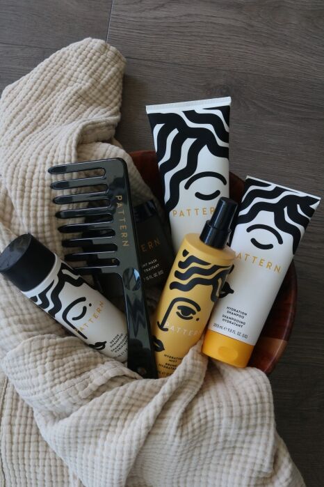 A bowl with Pattern hair products—shampoo, conditioner, spray, gel—and a black wide-tooth comb, all on a beige textured fabric.