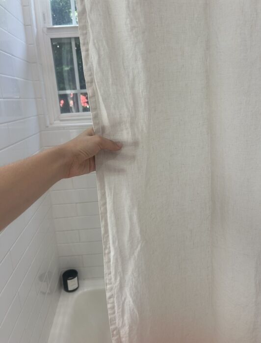 A hand holds a white shower curtain inside a tiled bathroom with a window and a black candle on the bathtub ledge.