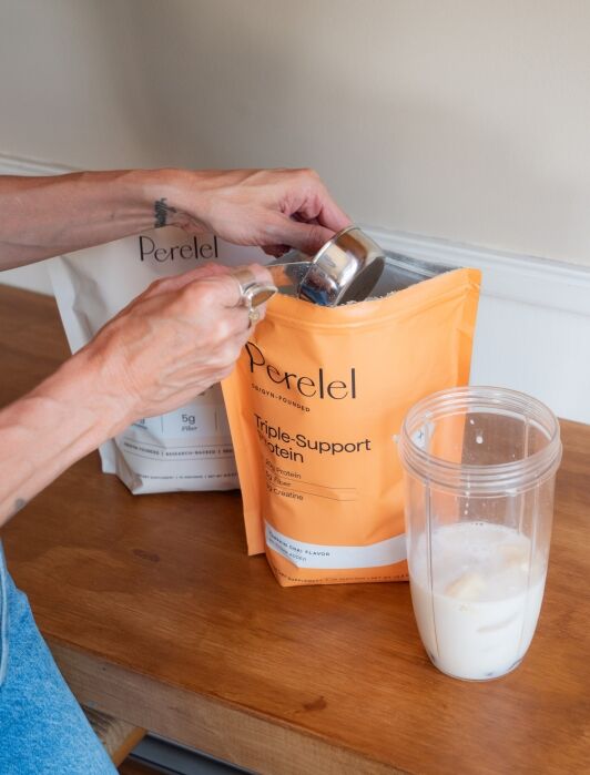 A person scoops protein powder from an orange Perel protein bag into a blender cup containing milk.