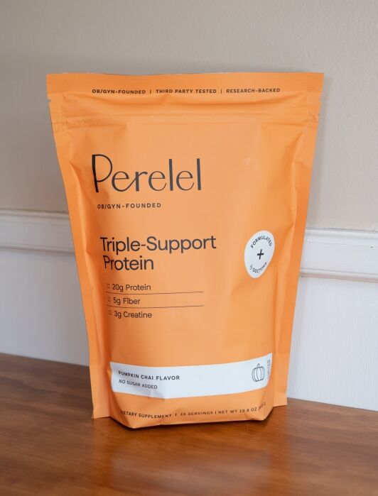 Orange bag of Perelel Triple-Support Protein powder in pumpkin chai flavor sits on a wooden surface with nutritional info and branding visible.