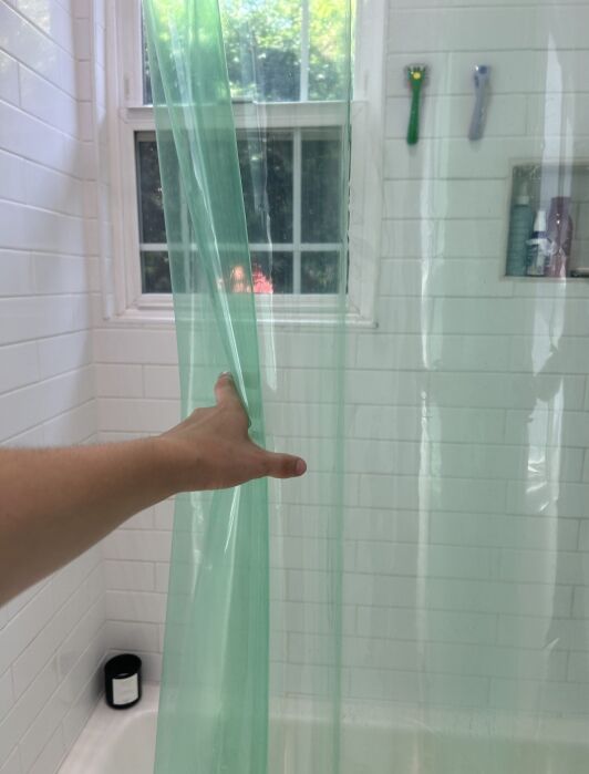 A hand pulls back a translucent green shower curtain in a white-tiled bathroom with a window and toiletries on a shelf.