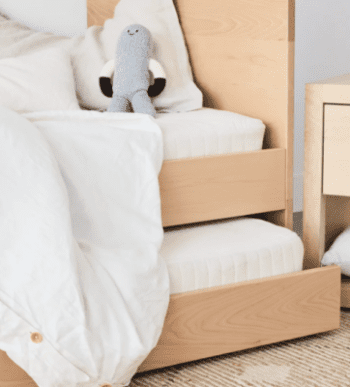Wooden trundle bed with white bedding and a knitted gray and white stuffed toy on top, next to a light wood nightstand.