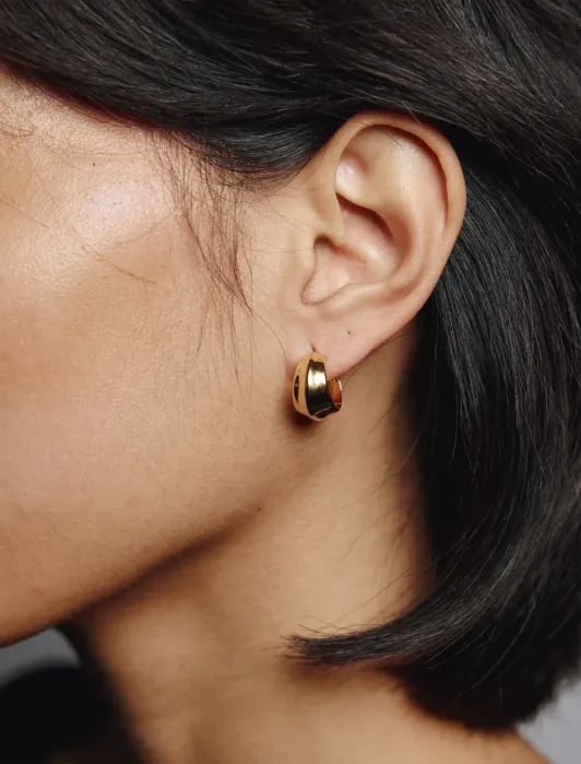 Close-up of a person with short dark hair wearing a small, thick gold hoop earring in their left ear.