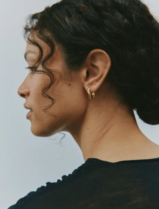 A woman with curly dark hair wears gold hoop earrings and a black top, shown in profile against a plain background.