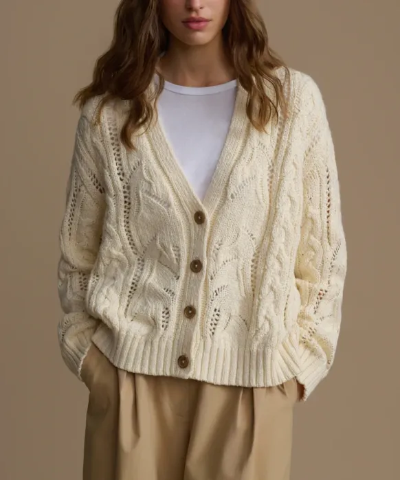 Woman wearing a cream cable-knit cardigan with brown buttons over a white shirt and tan pants, standing against a plain beige background.