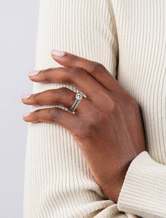 A person wearing a cream ribbed long-sleeve top rests their left hand on their right arm, showing two stacked rings with clear gemstones on their ring finger.
