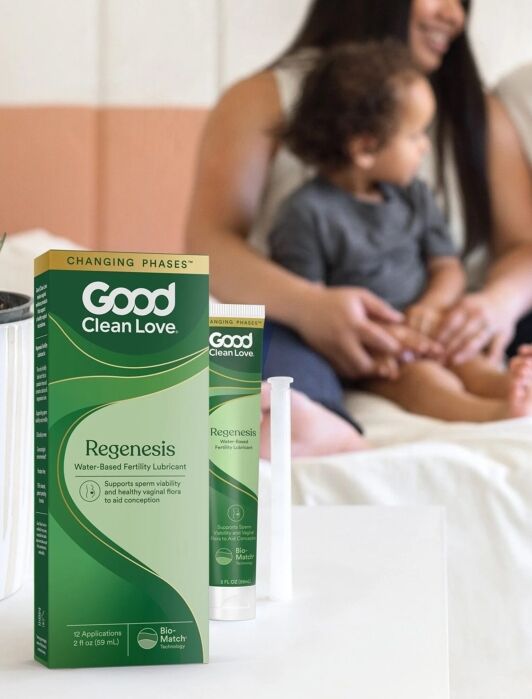 A box and tube of Good Clean Love fertility lubricant on a table with a blurred family of three sitting on a bed in the background.