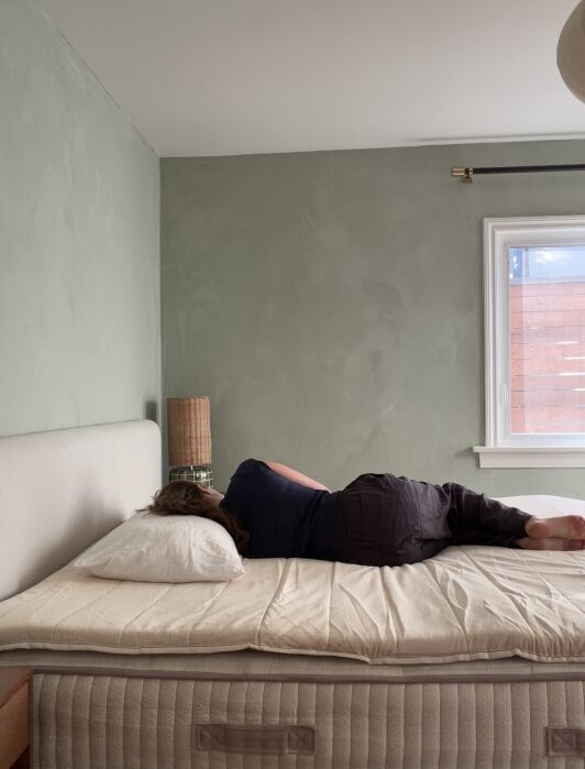 Person lying on their side on an unmade bed in a sparsely furnished bedroom with a light green wall and window.