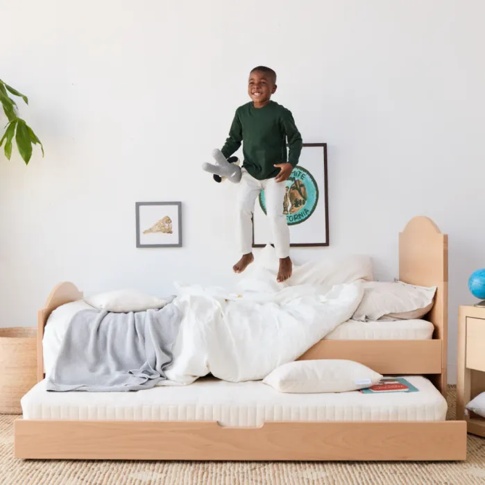 Smiling child jumps on an unmade light wood bed while holding a stuffed animal, surrounded by soft bedding and wall art in a bright, tidy room.