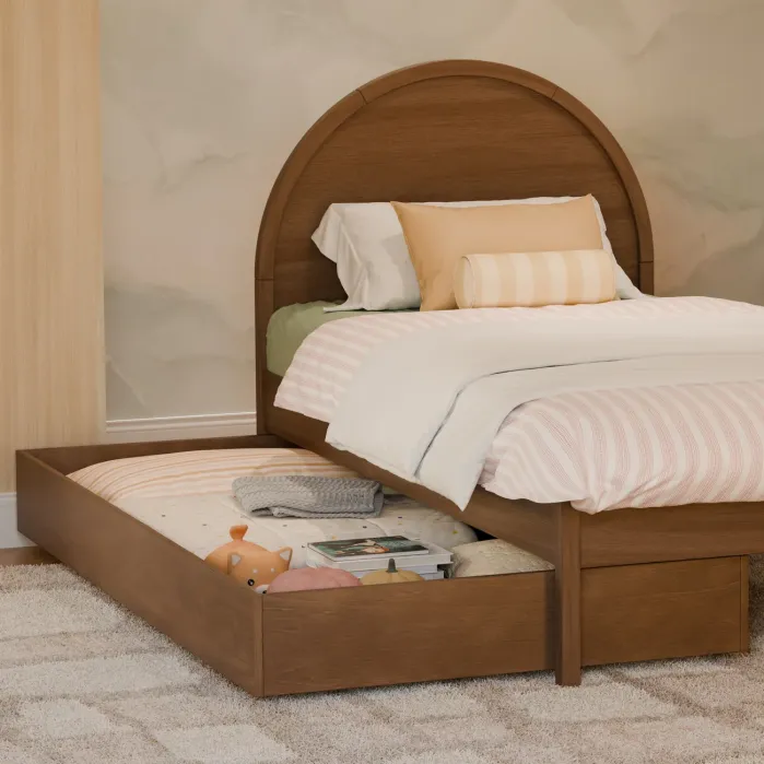 A wooden bed with a curved headboard features a pull-out trundle drawer containing a mattress, books, and a teddy bear, placed on a carpeted floor.