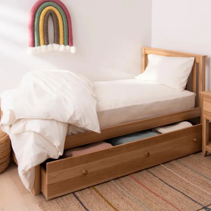 Single wooden bed with white bedding, a pull-out drawer underneath holding blankets, a bedside table, and a woven rainbow wall hanging above the bed.