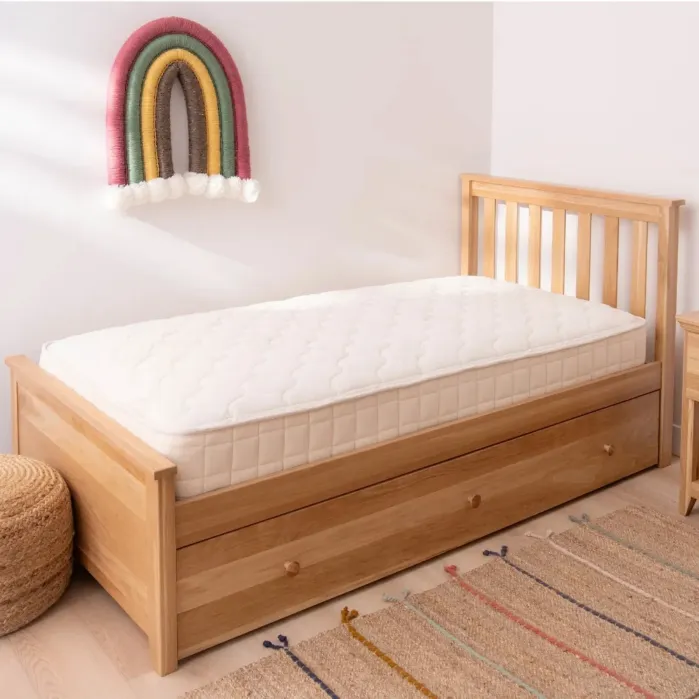 A wooden single bed with a white mattress, pull-out storage drawers, and a rainbow wall decoration above, set on a woven rug in a bright, simple room.